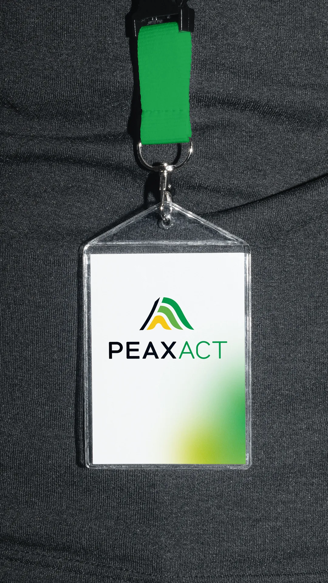 Namensschild mit grünem Band vor dunkelgrauem Shirt, darauf das neue PEAXACT Logo mit abstrahiertem Wellenmotiv in Gelb, Schwarz und Grün zur Veranschaulichung des Rebrandings. Gestaltung und Umsetzung durch die R211-Agentur