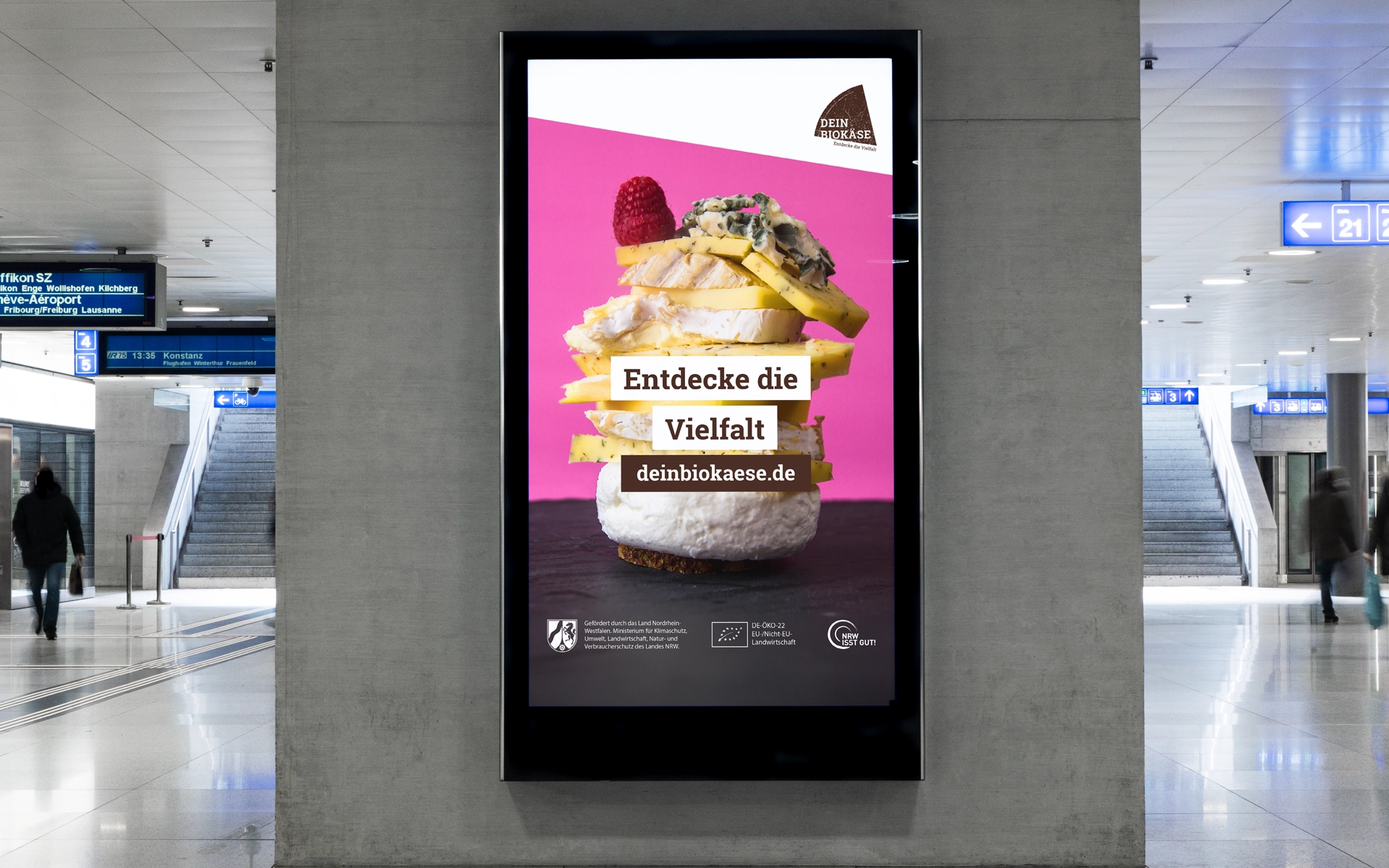 Digitales Plakat mit Käseprodukten auf pinkem Hintergrund in U-Bahn-Station, Text: Entdecke die Vielfalt deinbiokaese.de