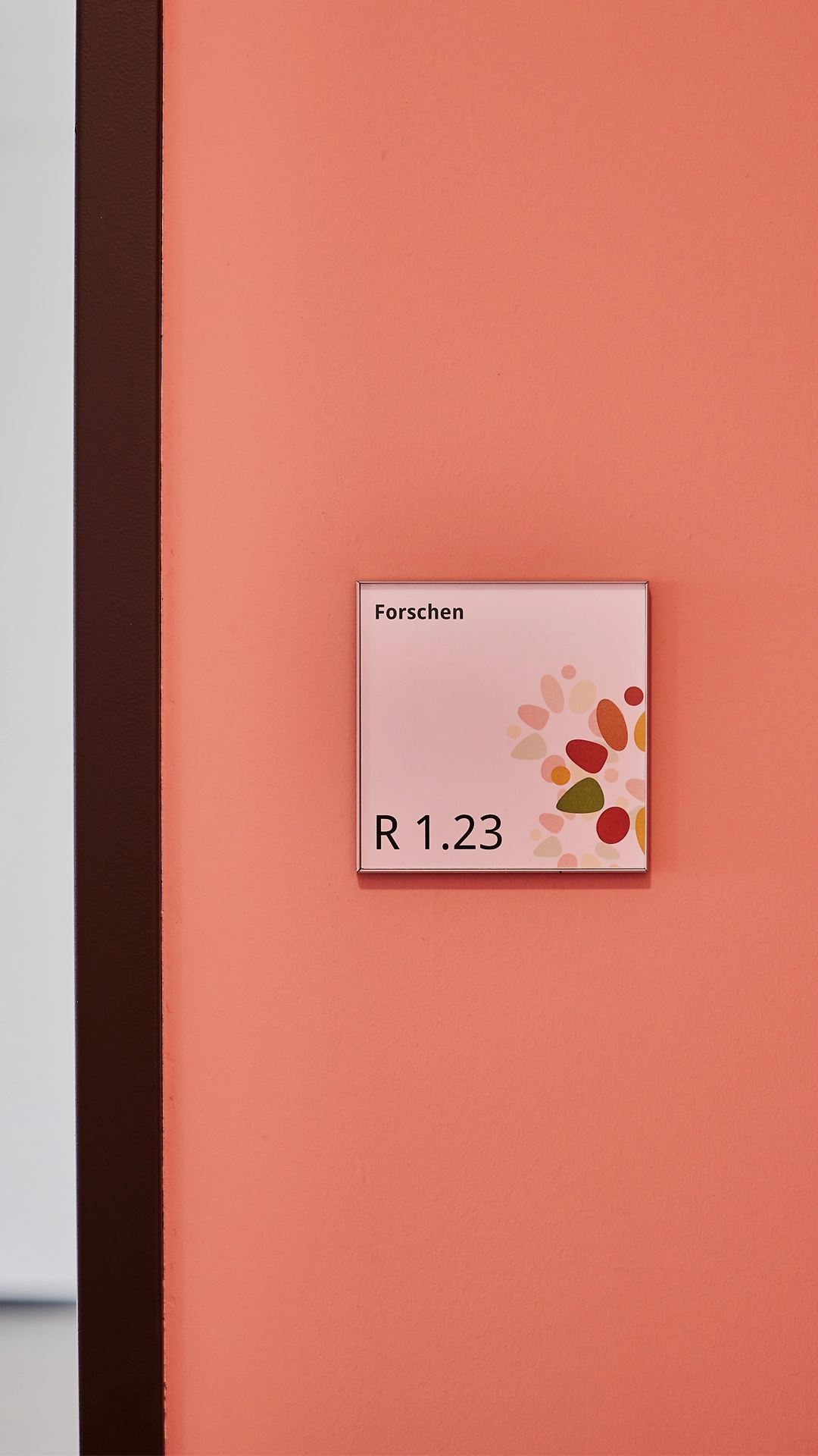 Türschild mit Raumbezeichnung „Forschen – R 1.23“ auf rosafarbener Wand in der Grundschule „Im Pfingsterfeld“, gestaltet von R211 als Teil des farbcodierten Leitsystems mit organischem Motiv