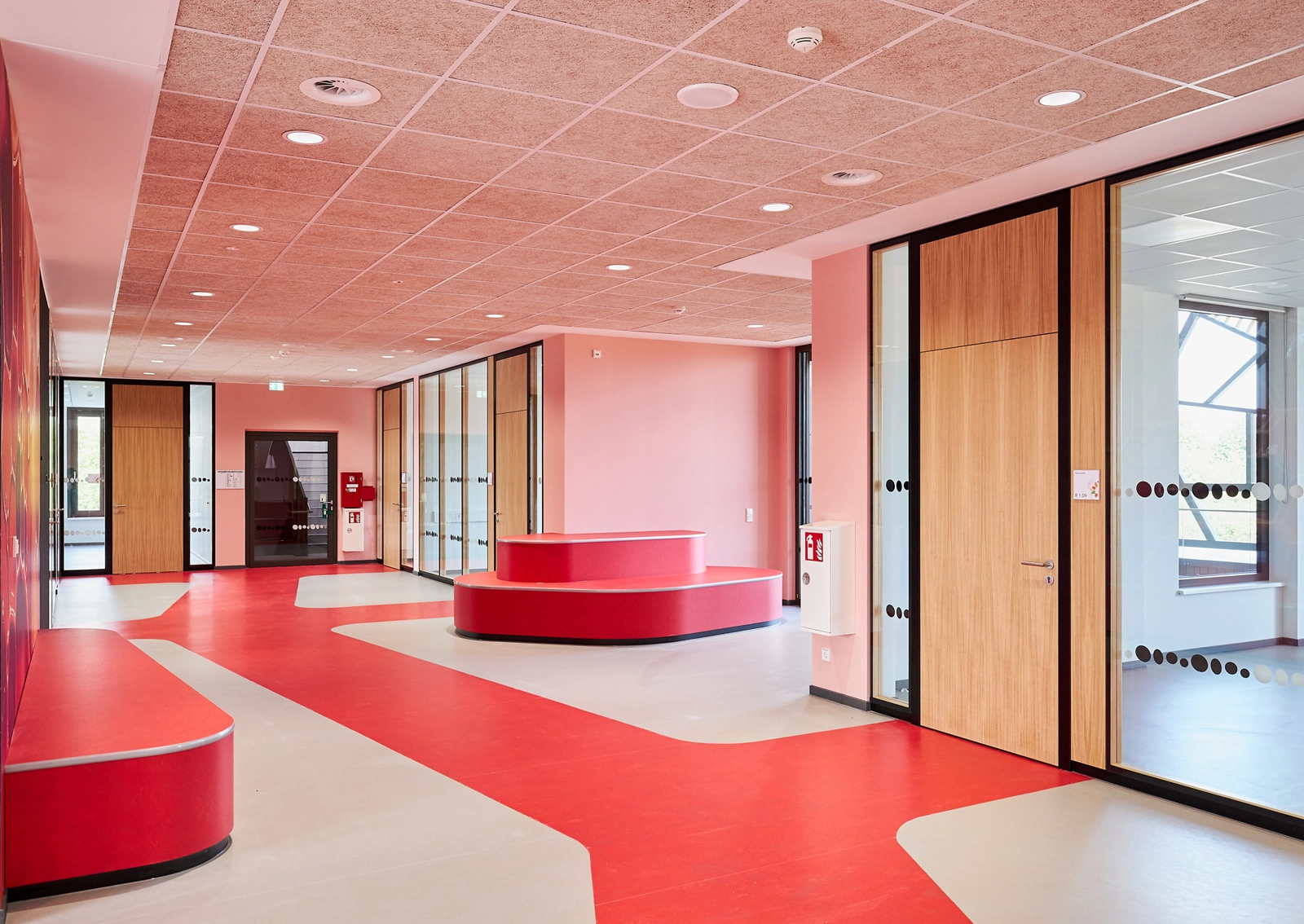 Innenraum der Grundschule „Im Pfingsterfeld“ mit rot akzentuiertem Clusterbereich, Sitzmöbeln und Glastüren mit Punktmarkierung – Teil des farbcodierten Leitsystems von R211 zur Unterstützung eines modernen pädagogischen Konzepts