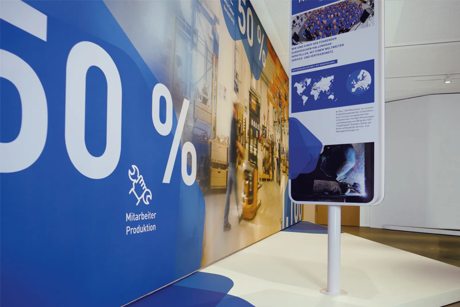 Kommunikationskonzept eines Showrooms für ABUS Kransysteme mit Ausstellungswand mit blauer Gestaltung zur Produktion und Infostele mit Weltkarte und Unternehmensdaten